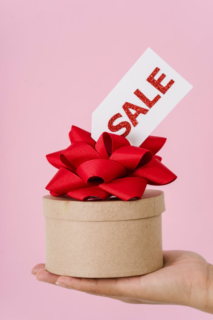 A close-up of a hand holding a round gift box with a red bow and sale tag on a pink background.